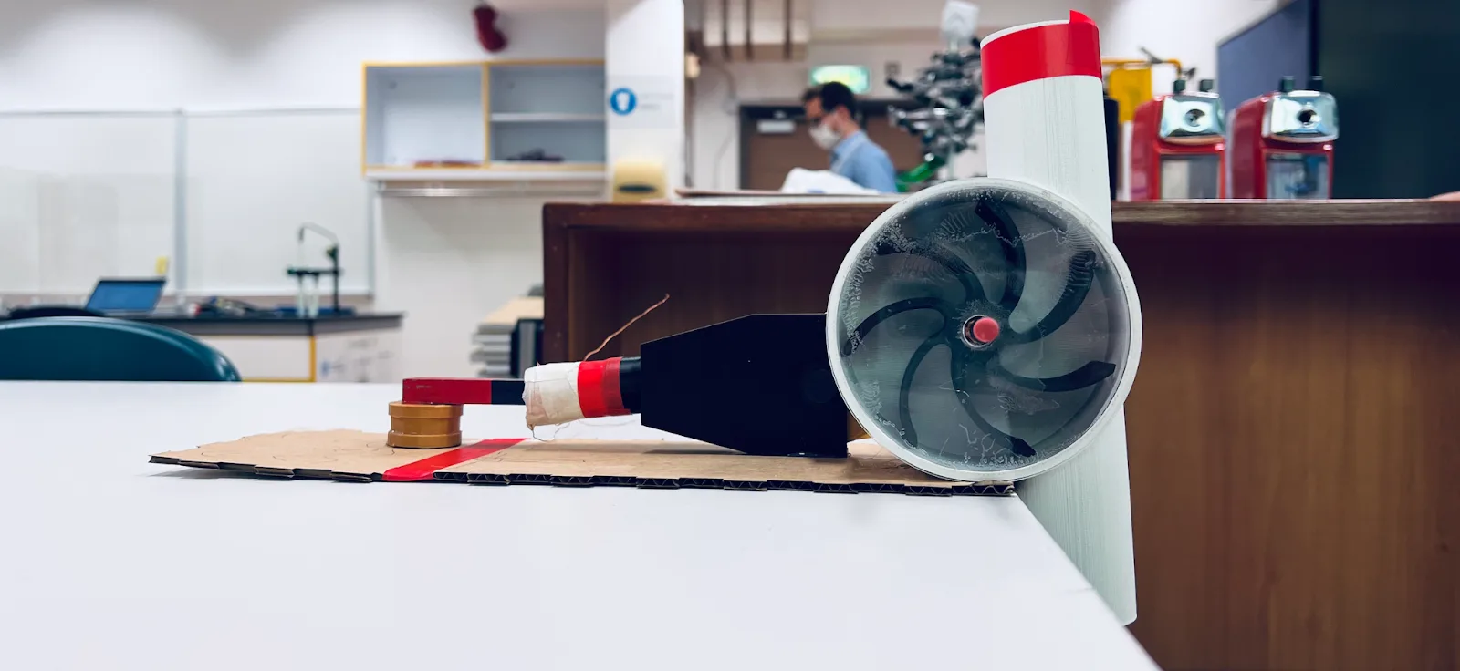 The working FLOW prototype on a classroom bench: a white 3D-printed pipe section with a clear-faced turbine housing showing curved black waterwheel blades, connected through a black housing to a red-and-white magnet and copper-wire coil assembly on a cardboard base