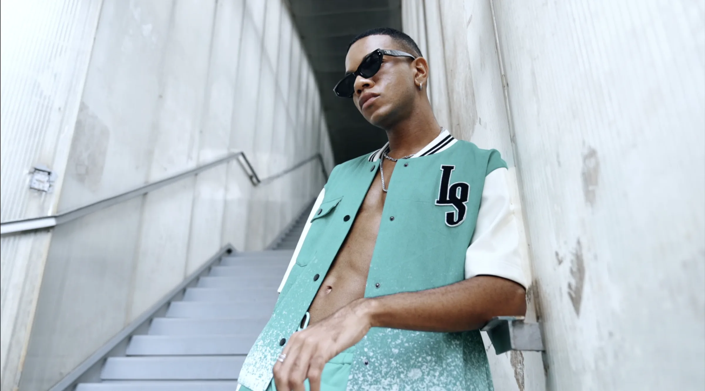 Low-angle shot of a model leaning against a concrete stairwell wall in the cyan-and-cream Chip Body bomber with an embroidered LS monogram and black sunglasses